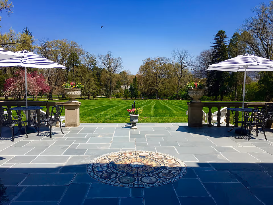 Stone patio with tables and striped umbrellas overlooking a manicured lawn and trees under a clear blue sky.
