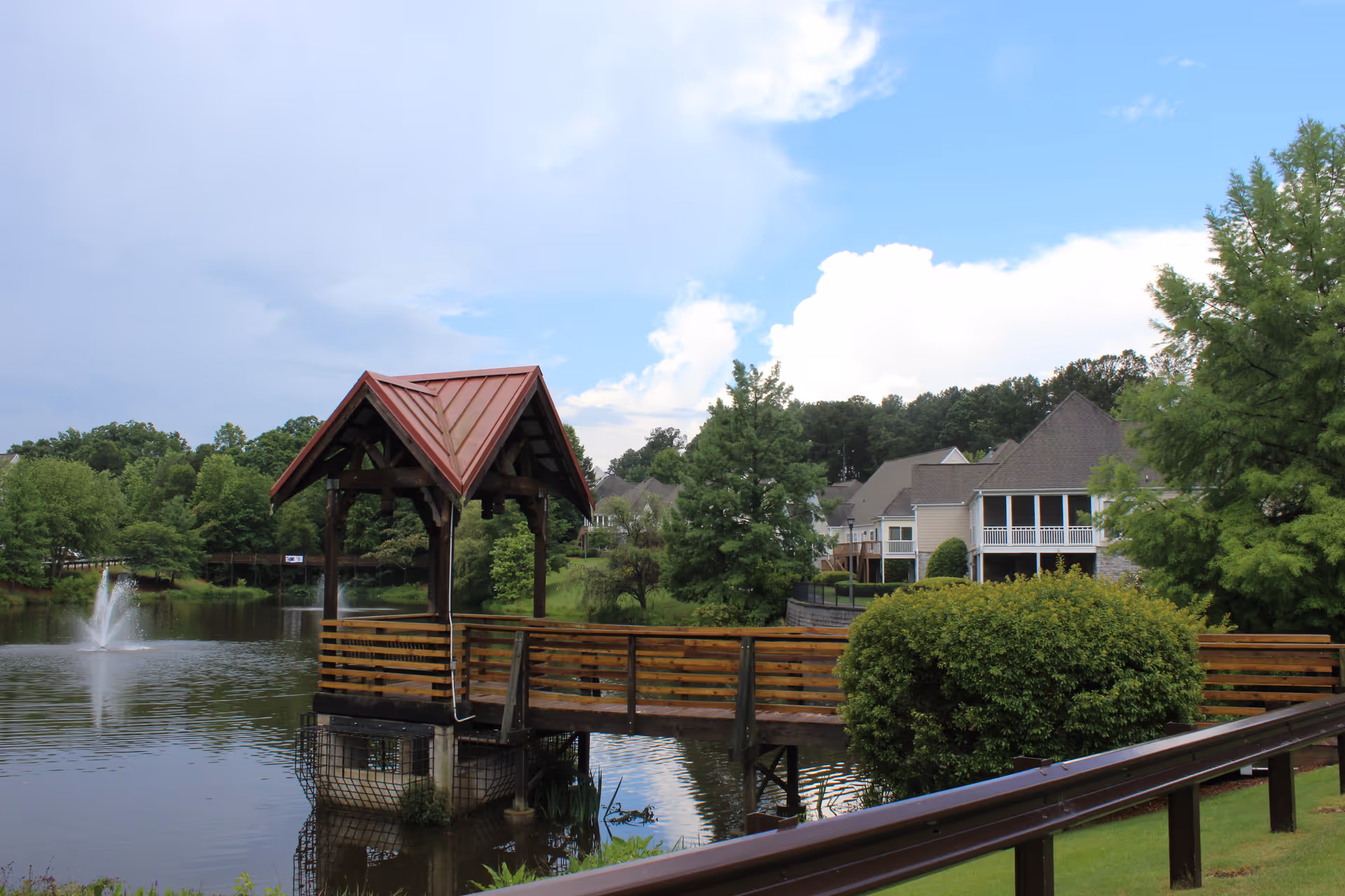 A wooden gazebo with a red roof on a pier extending over a pond with a water fountain. Surrounding the pond are green trees and shrubs, with residential buildings visible in the background under a partly cloudy sky.