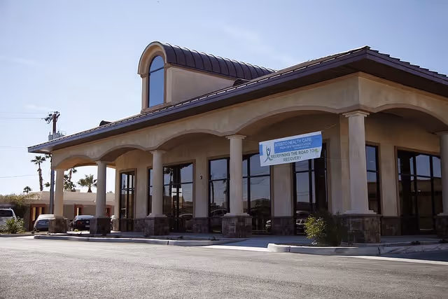 Exterior view of Kissito Healthcare - Palm View building with a covered entrance supported by columns and a sign that reads 'Kissito Health Care Supporting the Road to Recovery'. Several vehicles are parked nearby under a clear sky.