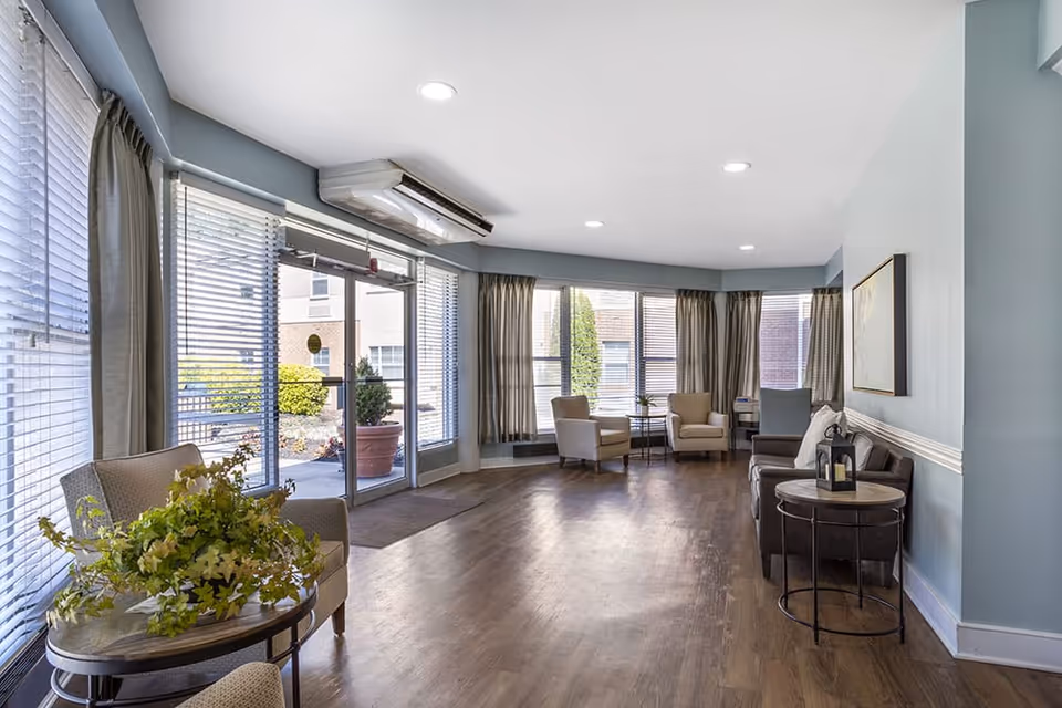 A bright and spacious seating area in a senior living facility with large windows covered by blinds and curtains. The room features wooden flooring, several armchairs, a sofa, small round tables with decorative items, and a glass door leading outside. The walls are painted light blue with white trim, and recessed ceiling lights illuminate the space.