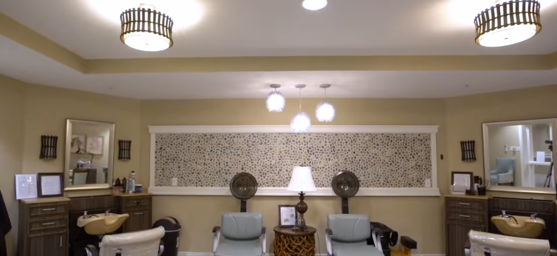 Interior view of a salon area in an assisted living facility featuring two salon chairs with sinks, two light blue chairs, a small round table with a lamp, two large mirrors on the walls, and decorative wall art with pebble-like texture. The room is softly lit with ceiling lights and pendant lights.