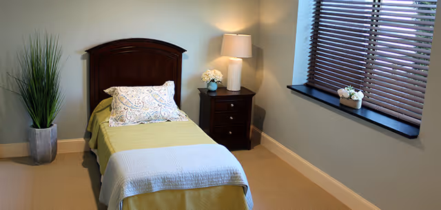 A small, neatly arranged bedroom with a single bed covered in a green and light blue blanket, a decorative pillow, a wooden headboard, a nightstand with a lamp and a small flower vase, a tall potted plant, and a window with wooden blinds and a small flower pot on the windowsill.