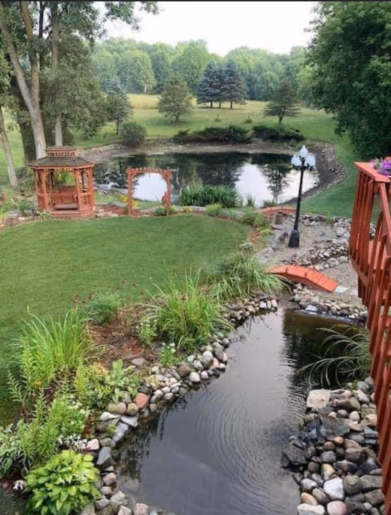 Landscaped backyard with rock-lined ponds, a wooden gazebo, a small arched bridge and surrounding trees.