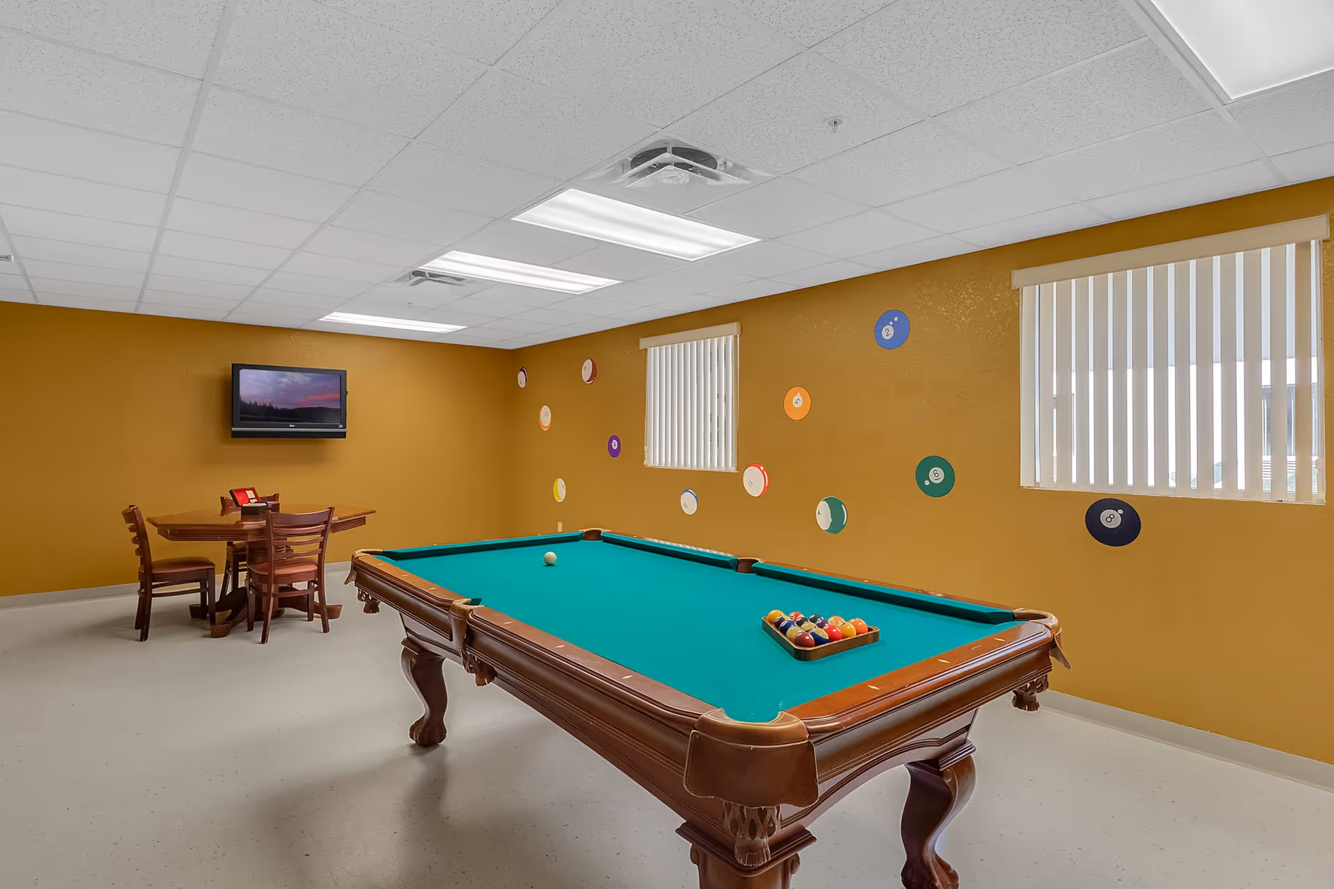 A recreational room with a pool table in the center, a small table with four chairs in the corner, a wall-mounted TV, and yellow walls decorated with colorful billiard ball designs. The room has two windows with vertical blinds and a white ceiling with fluorescent lights.