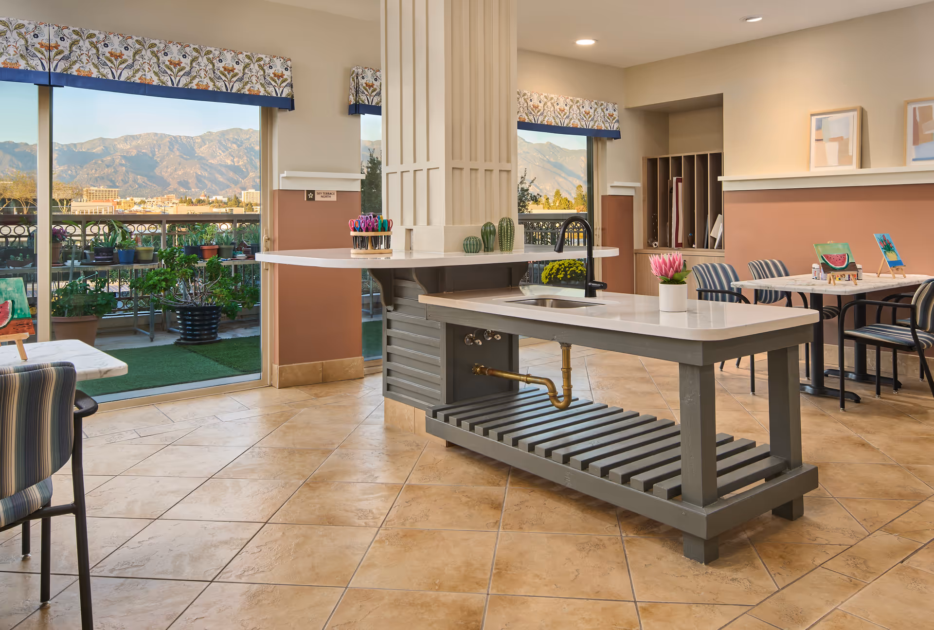 A bright and spacious common area in a senior living facility featuring a central island with a sink and decorative plants. The room has tiled floors, several tables and chairs with striped upholstery, and large windows with floral valances that offer a view of mountains and an outdoor garden area with potted plants.