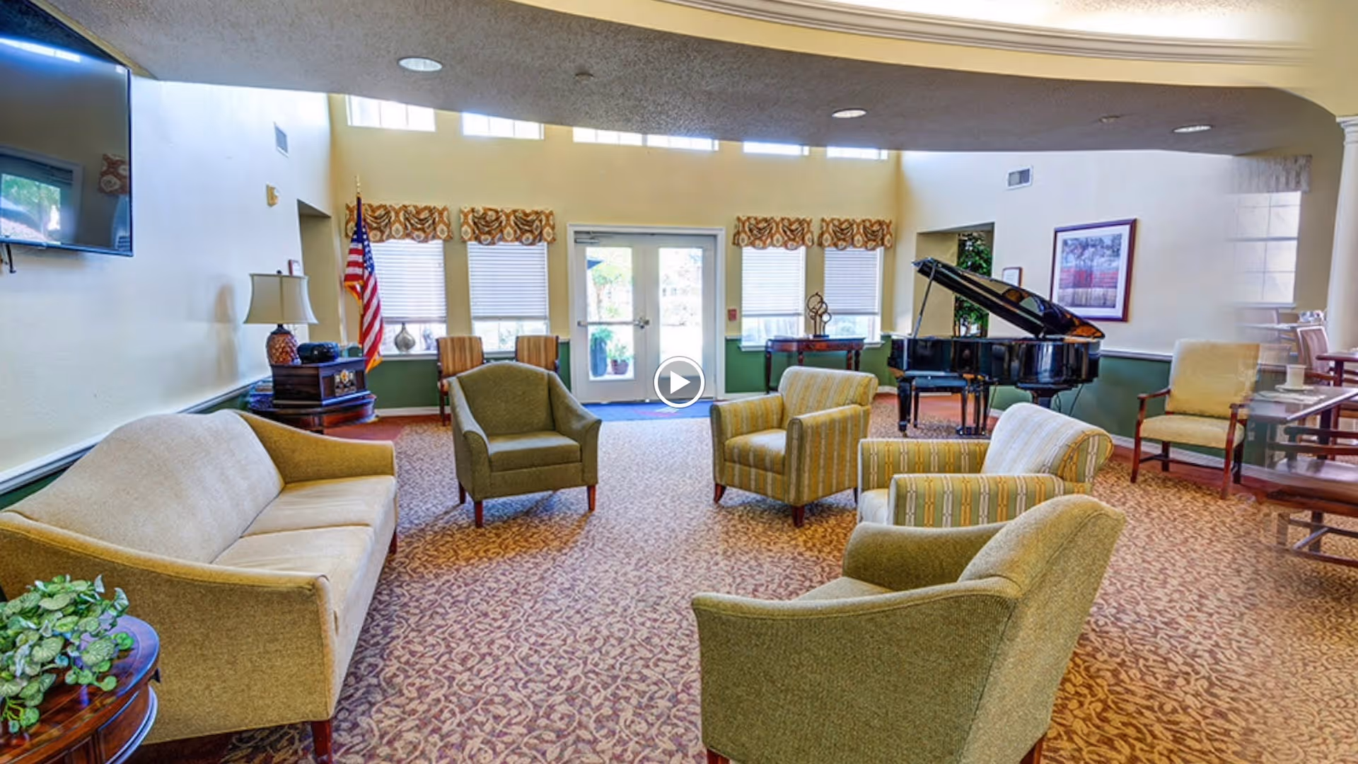 Bright common lounge with upholstered chairs and couches arranged around a grand piano near windows and an American flag.