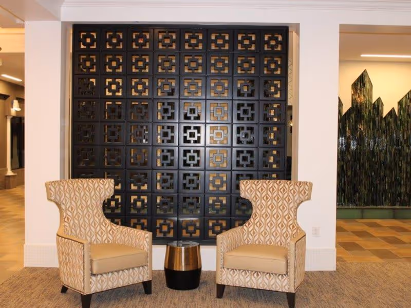 Two patterned armchairs with beige cushions are positioned on either side of a small round black and gold table in a room with a decorative black partition wall behind them. The floor is carpeted and the walls are white, with a glimpse of another room featuring a green textured wall visible through an opening on the right.