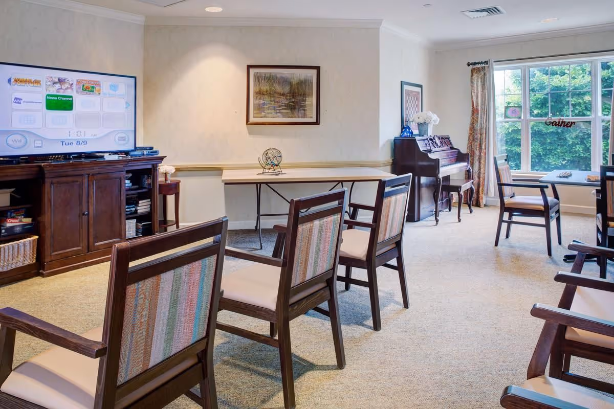 A well-lit common room with several wooden chairs arranged around tables. There is a large flat-screen TV on a wooden cabinet displaying a menu screen, a framed painting on the wall, and a piano near a window with floral curtains. The room has carpeted flooring and a bright window showing greenery outside.