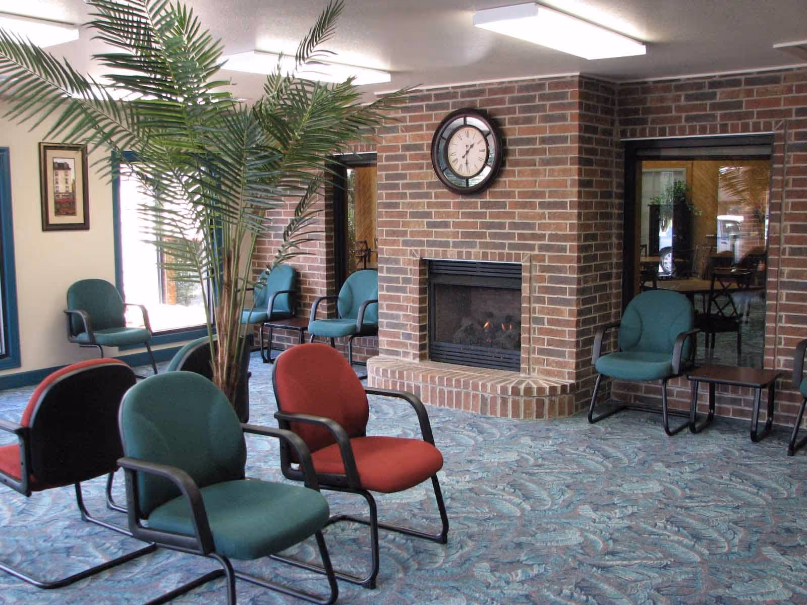 A cozy common area with a brick fireplace, a clock above it, several green and red cushioned chairs arranged around the room, a large potted plant, and a patterned blue carpet.