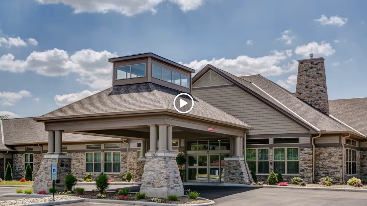 Exterior view of Sanctuary Pointe Nursing & Rehab Center showing the main entrance with a covered drop-off area supported by stone pillars, large windows, and a stone chimney under a partly cloudy sky.
