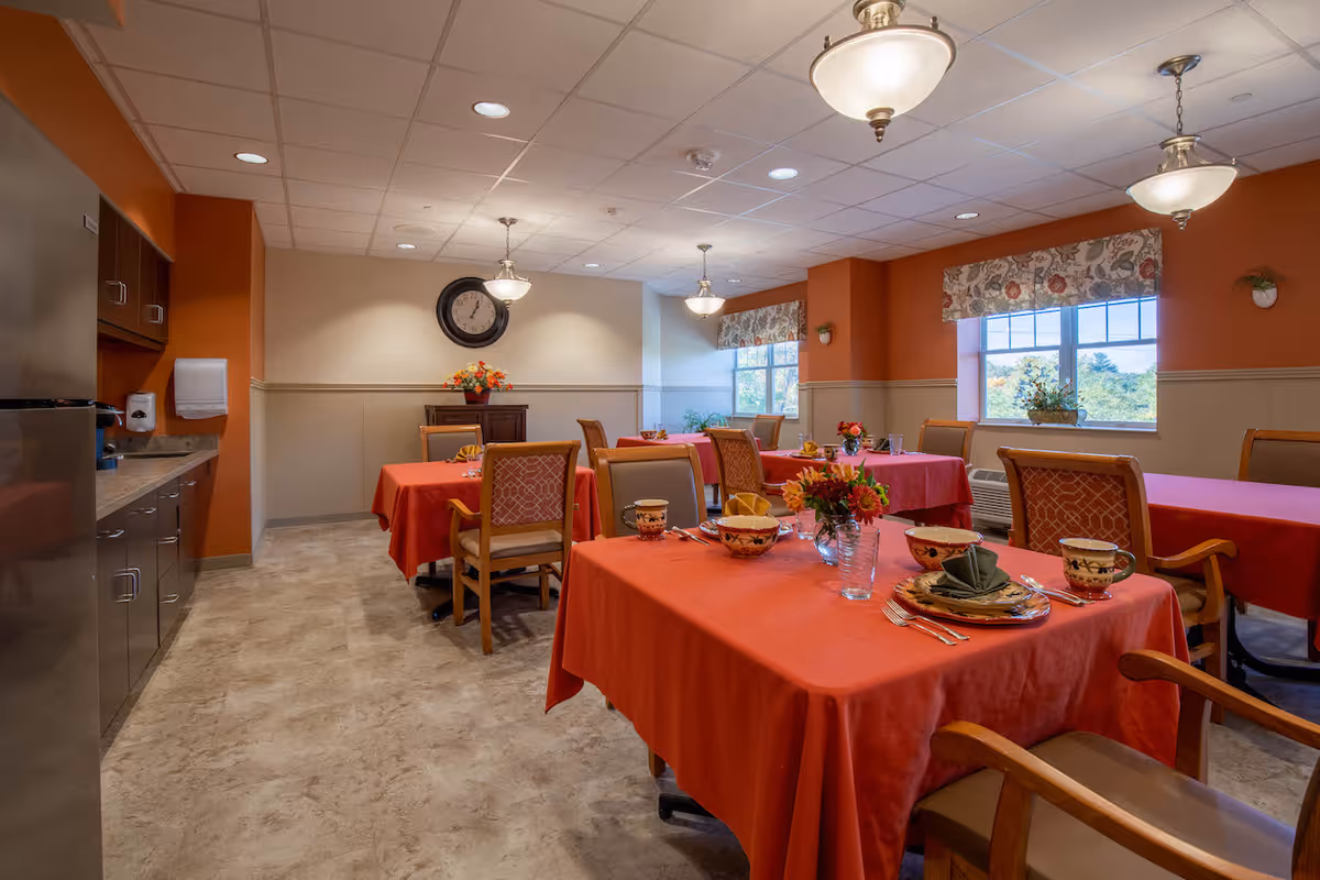 A dining room in Linwood Nursing and Rehabilitation Center with several tables covered in red tablecloths, each set with plates, cups, and utensils. The room has warm orange and beige walls, large windows with floral valances, ceiling lights, and a clock on the far wall. There is a kitchenette area with cabinets and a countertop on the left side.