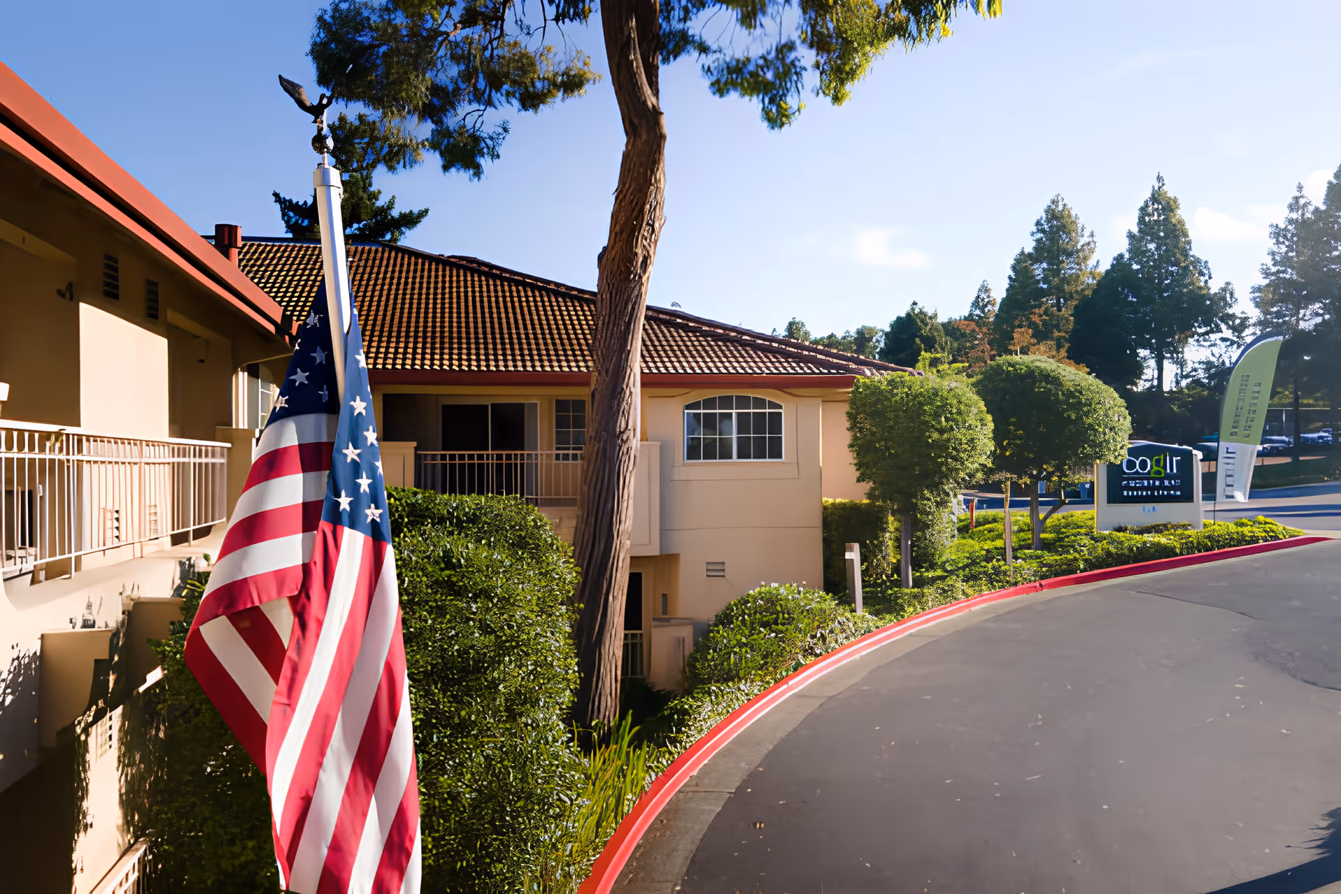 Exterior view of Cogir Of North Bay facility with an American flag in the foreground, a curved driveway bordered by red-painted curb, manicured bushes, trees, and a sign displaying the facility's name.