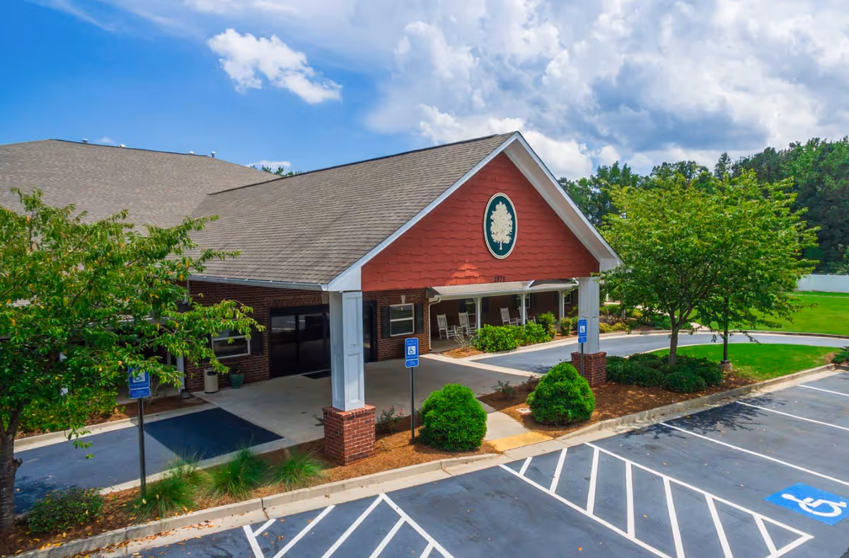 Exterior view of Oaks at Post Road facility entrance with a covered drop-off area supported by white columns and brick bases. The building has a red gable with a tree emblem, surrounded by green bushes and trees. There are several handicap parking spaces in front, and the sky is partly cloudy.