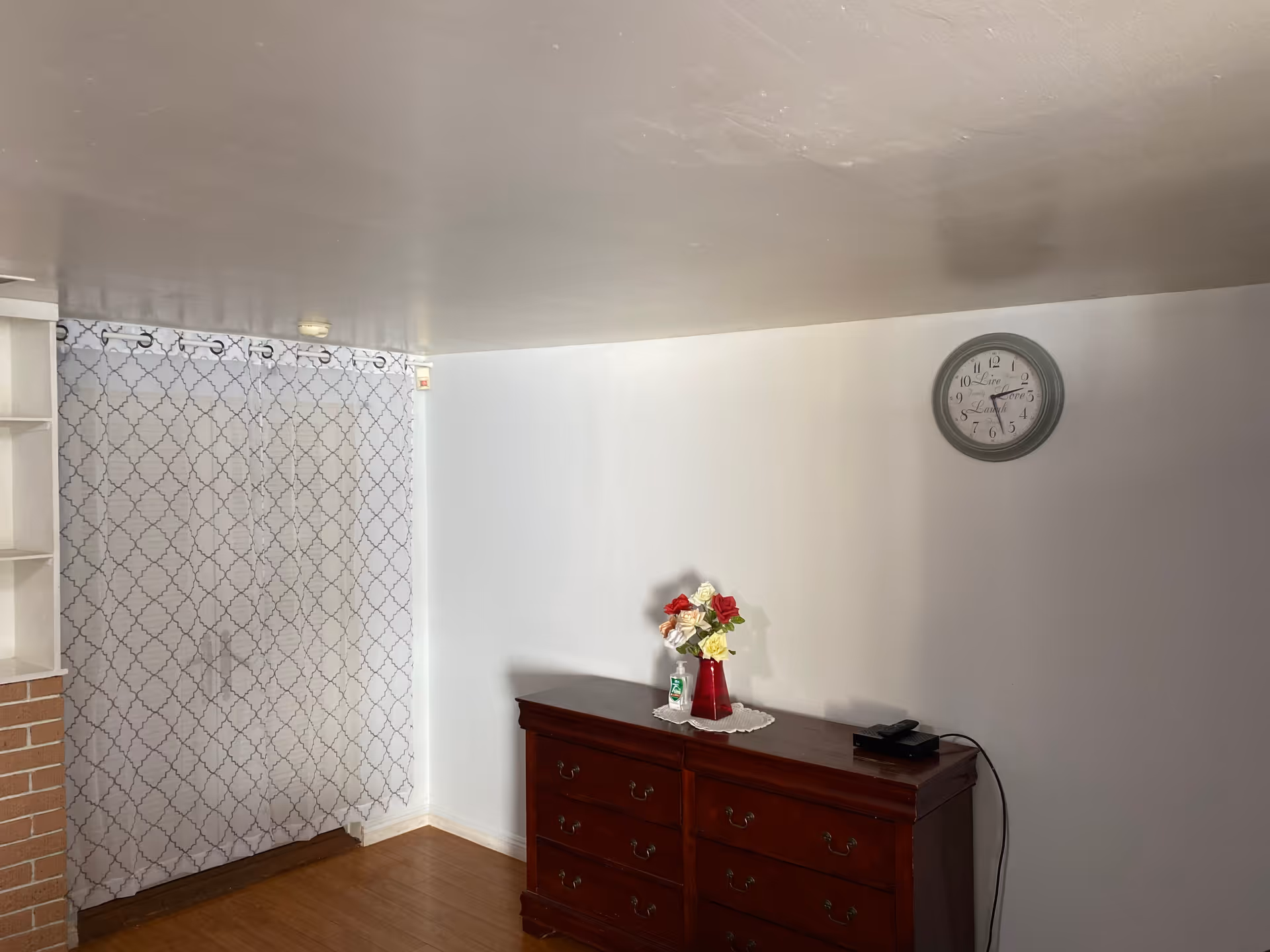 A room with a wooden dresser against a white wall. On top of the dresser is a red vase with artificial flowers and a bottle of hand sanitizer. A round wall clock hangs above the dresser. To the left, there is a window covered with a white curtain featuring a geometric pattern. The floor is wooden.
