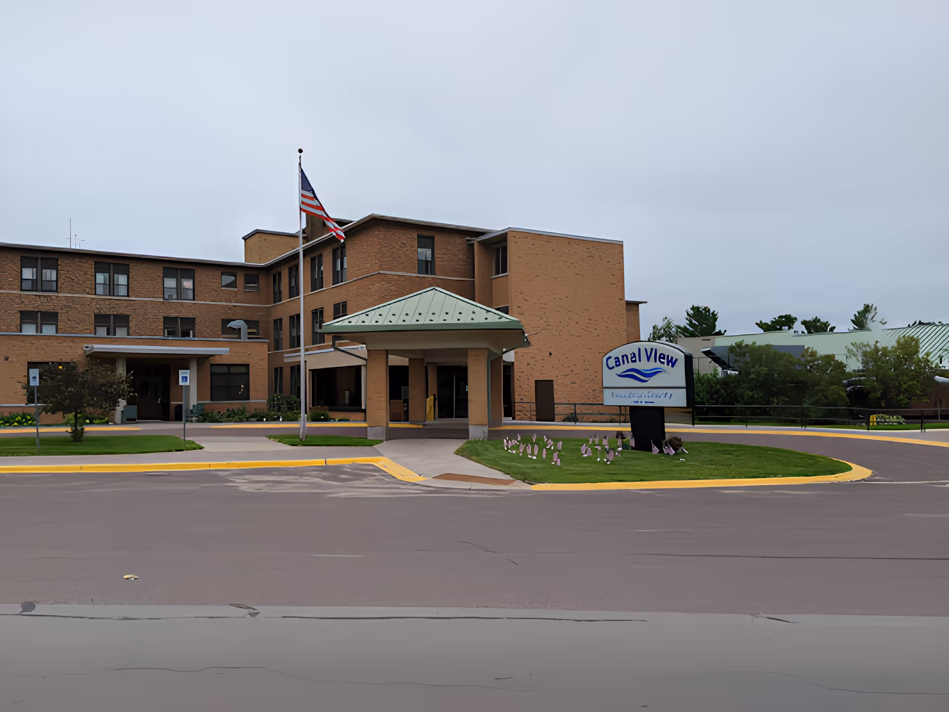 Exterior view of Canal View - Houghton County facility showing a three-story brick building with a covered entrance, an American flag on a flagpole, and a sign with the facility name on a grassy area with small flags.