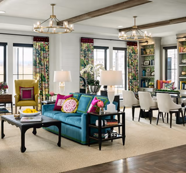 Bright open-plan living room with a teal sofa, colorful pillows, an armchair, coffee table, and a dining area in the background.
