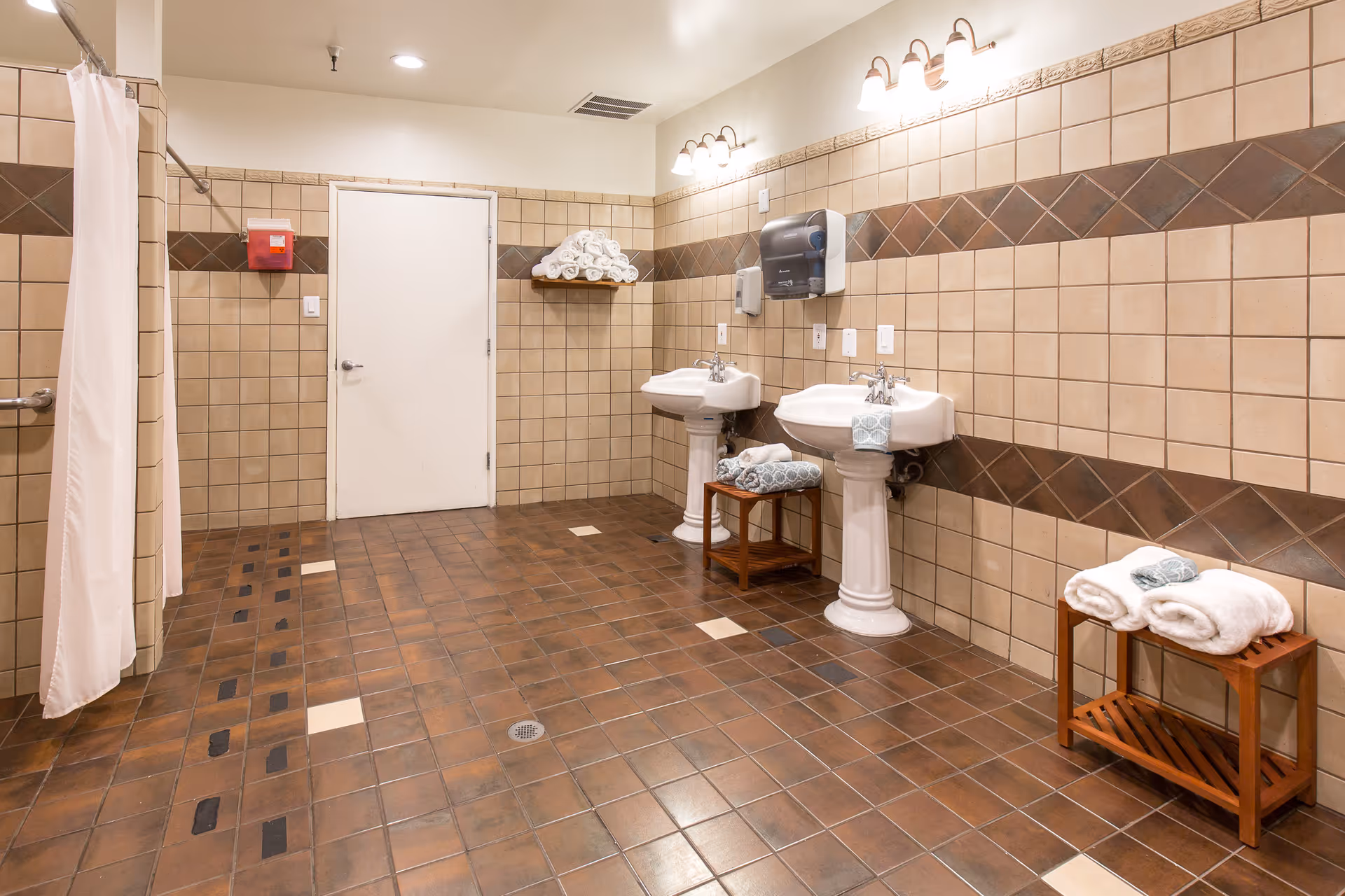 A spacious bathroom with brown and beige tiled walls and floor. The room features two white pedestal sinks with towels placed on small wooden stands beneath them. There is a white door in the center of the back wall, a shelf with rolled white towels above it, and a red wall-mounted container to the left. A shower area with a white curtain is visible on the left side of the image. The room is well-lit with ceiling lights and wall-mounted light fixtures above the sinks.