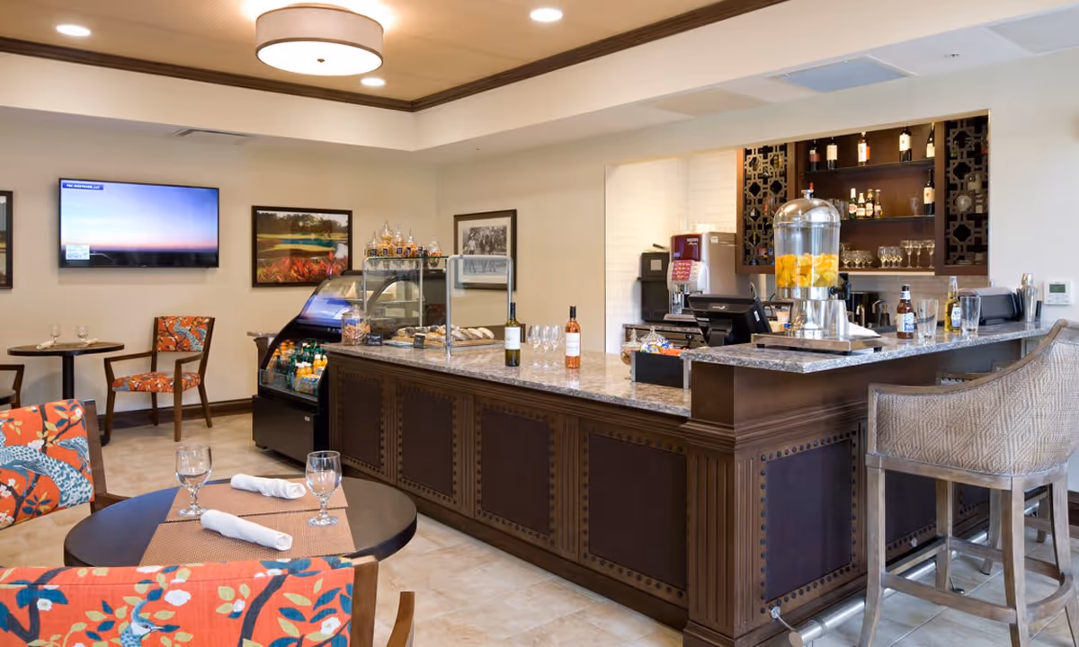 A cozy dining area with a granite countertop bar featuring a beverage dispenser with lemon water, bottles of wine, and glasses. There are small round tables with colorful floral-patterned chairs, set with placemats, napkins, and glasses. The walls have framed pictures and a flat-screen TV mounted. The space has warm lighting and a welcoming atmosphere.