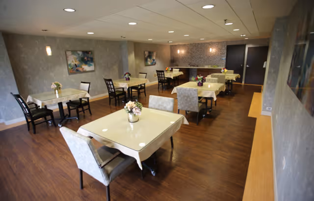A dining room with several square tables covered with light-colored tablecloths, each adorned with a small floral centerpiece. The room has wooden flooring, gray patterned walls, and modern ceiling lights. There are chairs arranged around the tables, and artwork is hung on the walls.