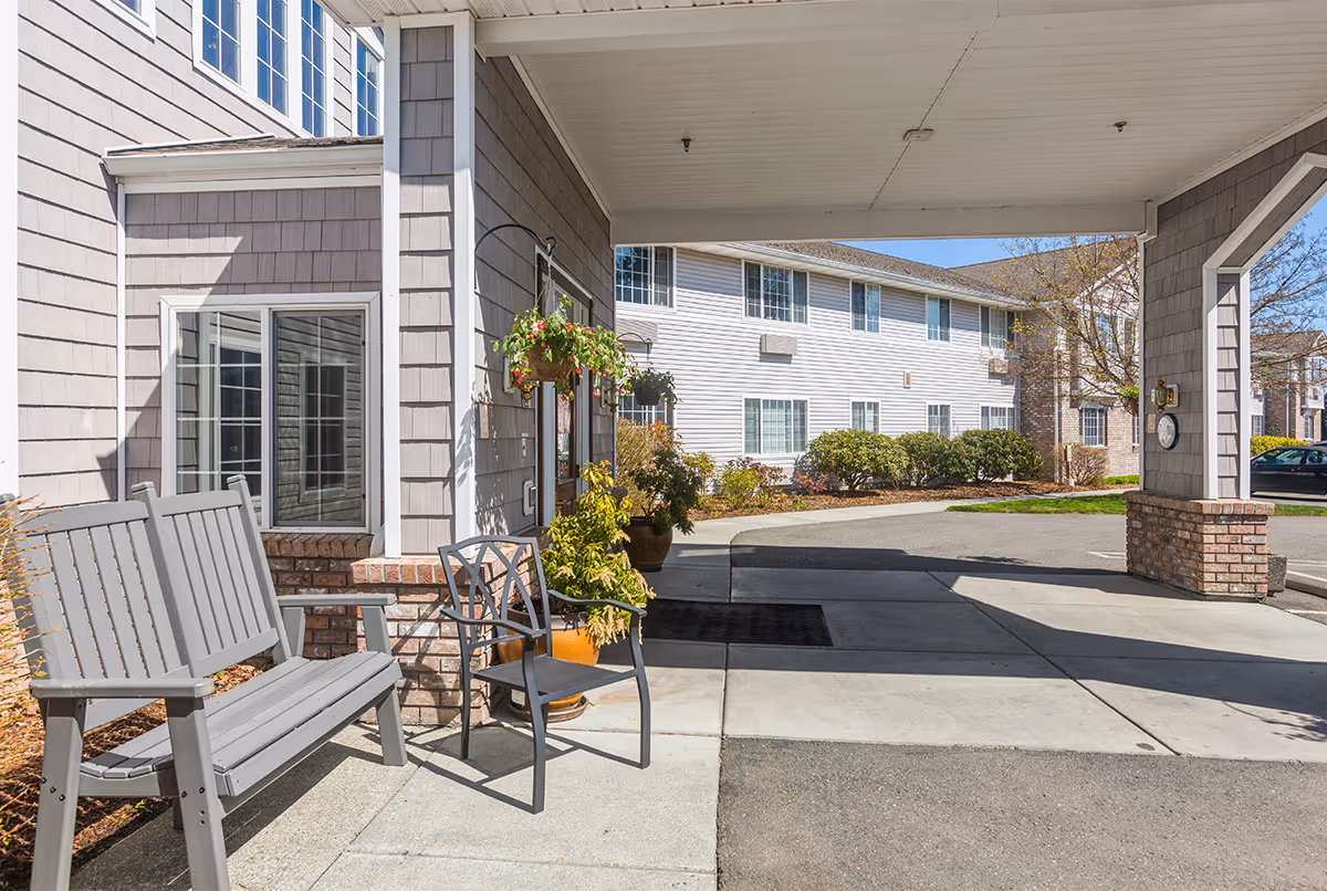 Covered entrance area of a senior living facility with a gray bench and a metal chair next to a potted plant and hanging flower basket. The building exterior features gray siding and white trim, with windows visible. A driveway and landscaped bushes are seen in the background under a clear blue sky.