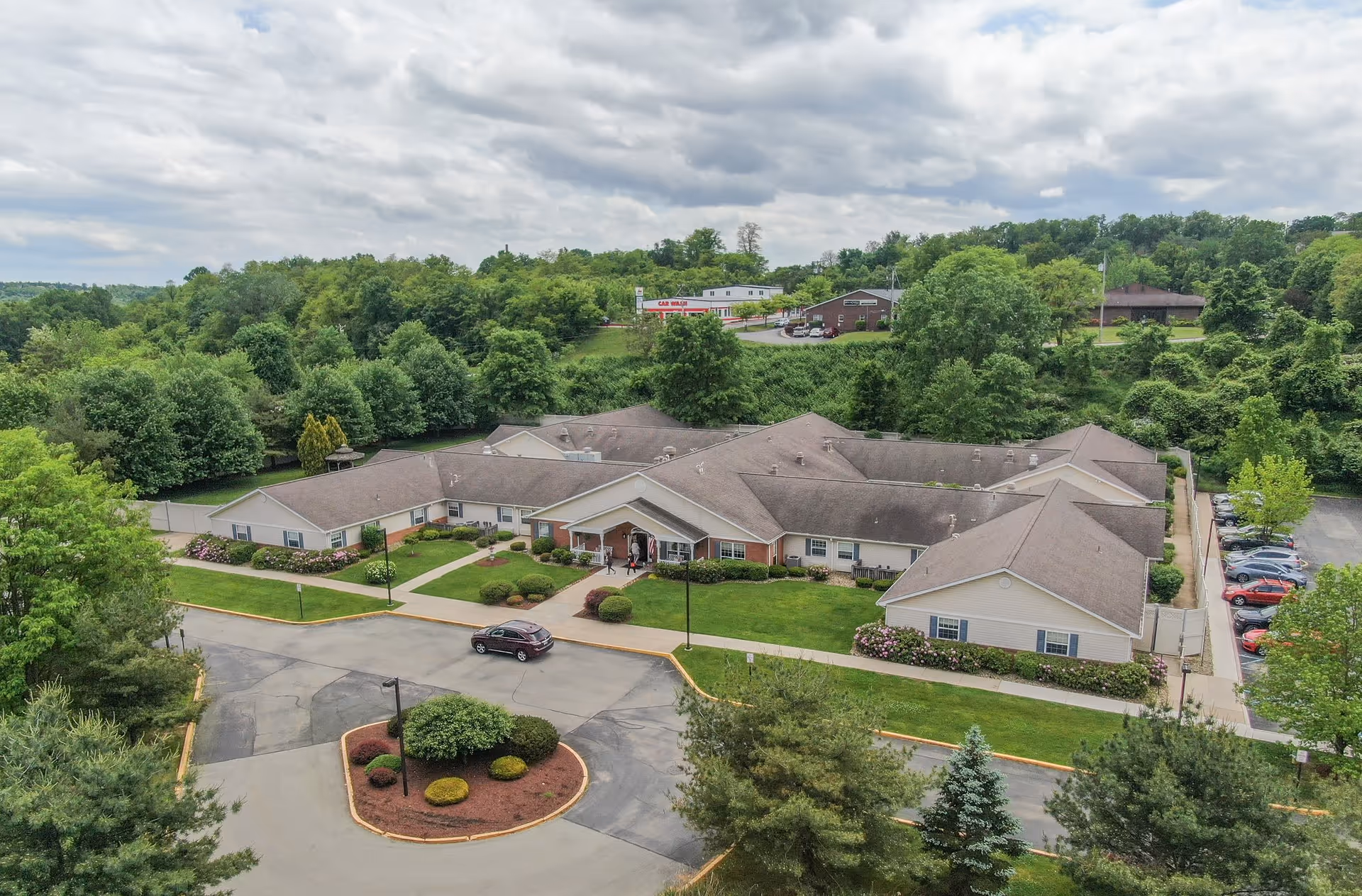 Aerial view of Arden Courts - ProMedica Memory Care Community in Monroeville, showing a single-story building surrounded by greenery, trees, and a parking area with several cars. The building has a light-colored exterior with a gray roof and landscaped lawns and bushes around it.