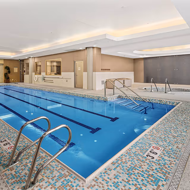 Indoor swimming pool with clear blue water, metal handrails, and tiled edges. The pool area features a hot tub in the background, beige walls, and a ceiling with recessed lighting.
