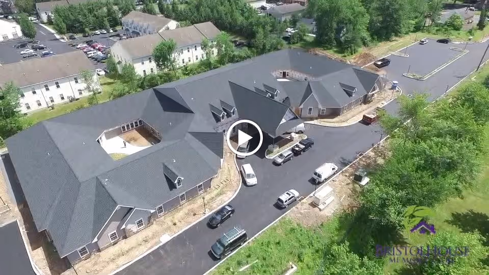 Aerial view of a large single-story building with a dark roof, surrounded by a newly paved driveway and parking area with several vehicles parked. The building is situated in a green area with trees and other residential buildings nearby. The bottom right corner shows a logo with the text 'Bristol House Memory Care'.