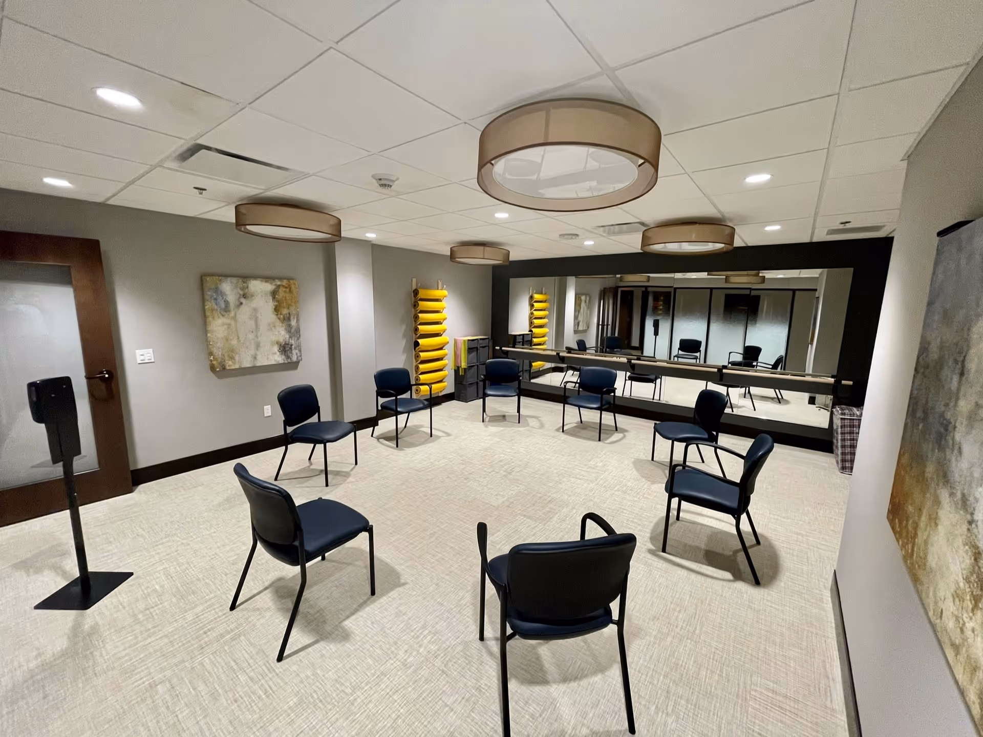 Well-lit interior activity room with chairs spaced in a circle, a wall-length mirror, stacked yellow exercise mats, and modern ceiling lights.