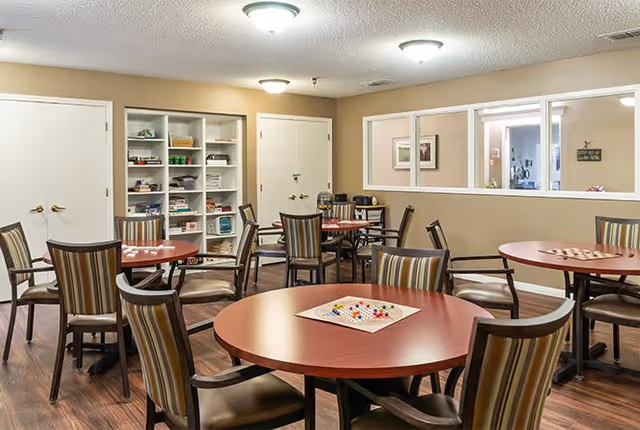 A well-lit common room with several round tables and striped cushioned chairs arranged around them. One table has a board game with colorful pieces set up. In the background, there is a white shelving unit filled with various games and items. The room has beige walls, wooden flooring, and a window looking into another room.