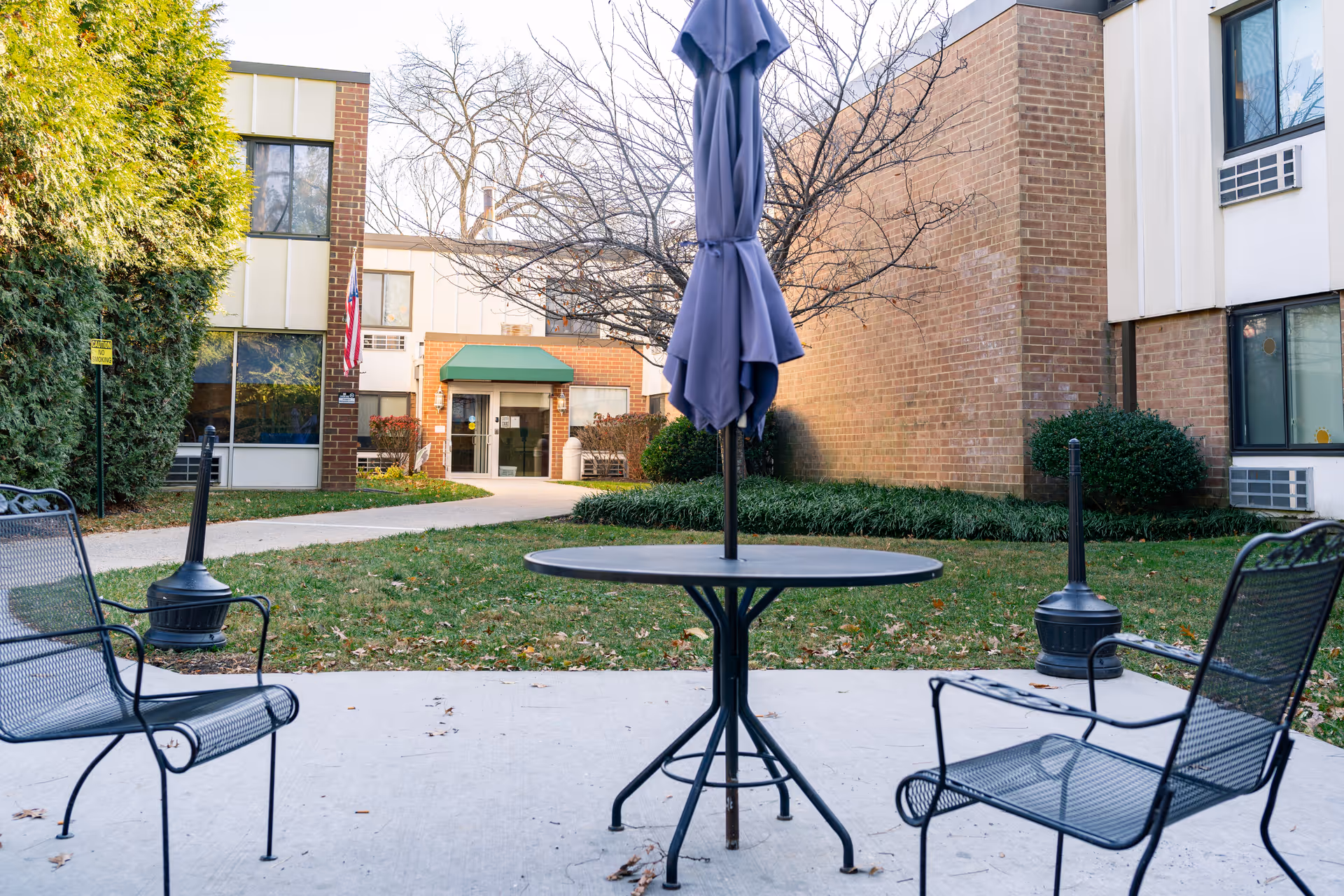 Outdoor seating area at Autumn Lake Healthcare at Catonsville featuring a round metal table with a closed umbrella in the center, surrounded by two metal chairs on a concrete patio. In the background, there is a pathway leading to the entrance of a brick and beige building with windows and an American flag near the door. Trees and bushes surround the area.