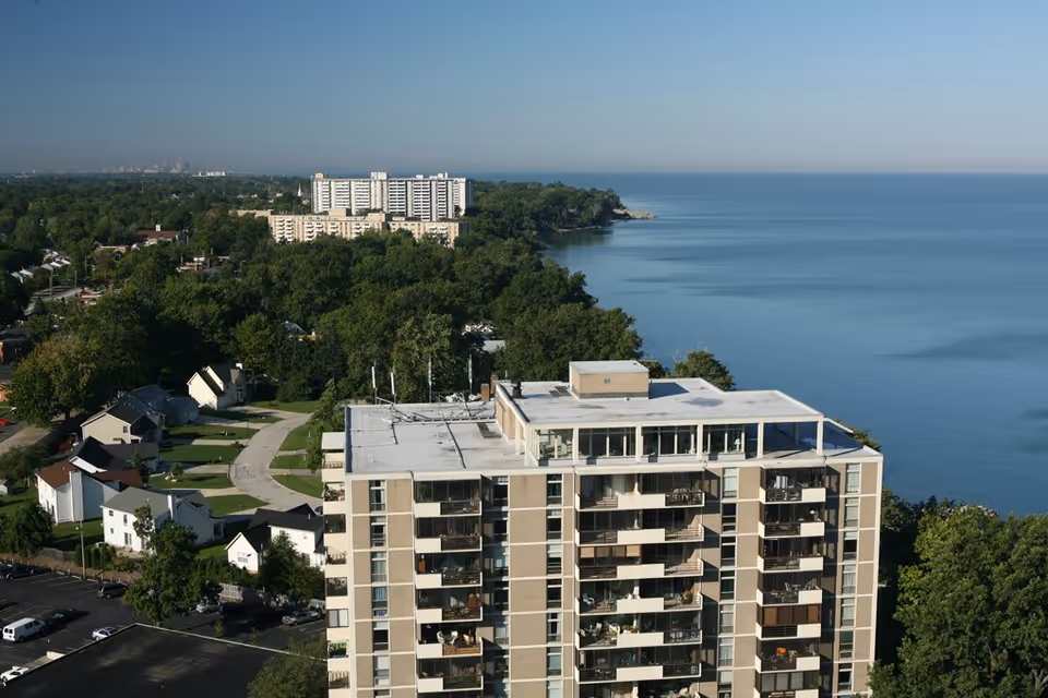 Aerial view of a multi-story residential building near a large body of water with a forested area and other buildings in the background under a clear blue sky.