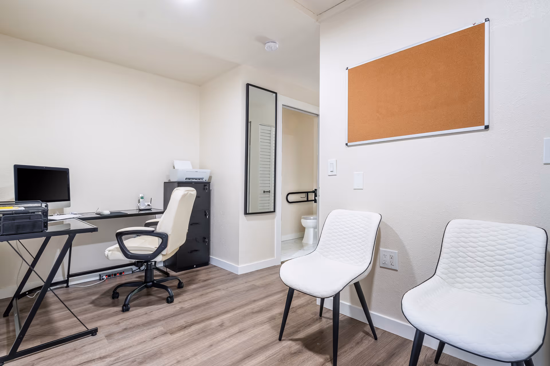 A small office area with a desk, computer, office chair, and filing cabinet. Two white chairs are positioned against the wall with a cork bulletin board above them. A doorway leads to a bathroom with a visible toilet and grab bar.