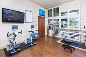 Small fitness room with two stationary bikes, an exercise ball, a weight rack, a wall-mounted TV, and large shuttered windows.