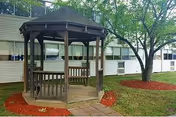 Wooden gazebo on a paved area surrounded by grass, red mulch beds, and trees in front of a low-rise building.