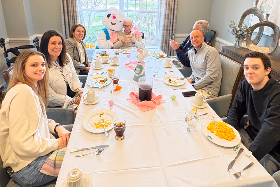 A group of people sit around a long dining table in a bright room sharing a meal, with an Easter bunny mascot at the far end.