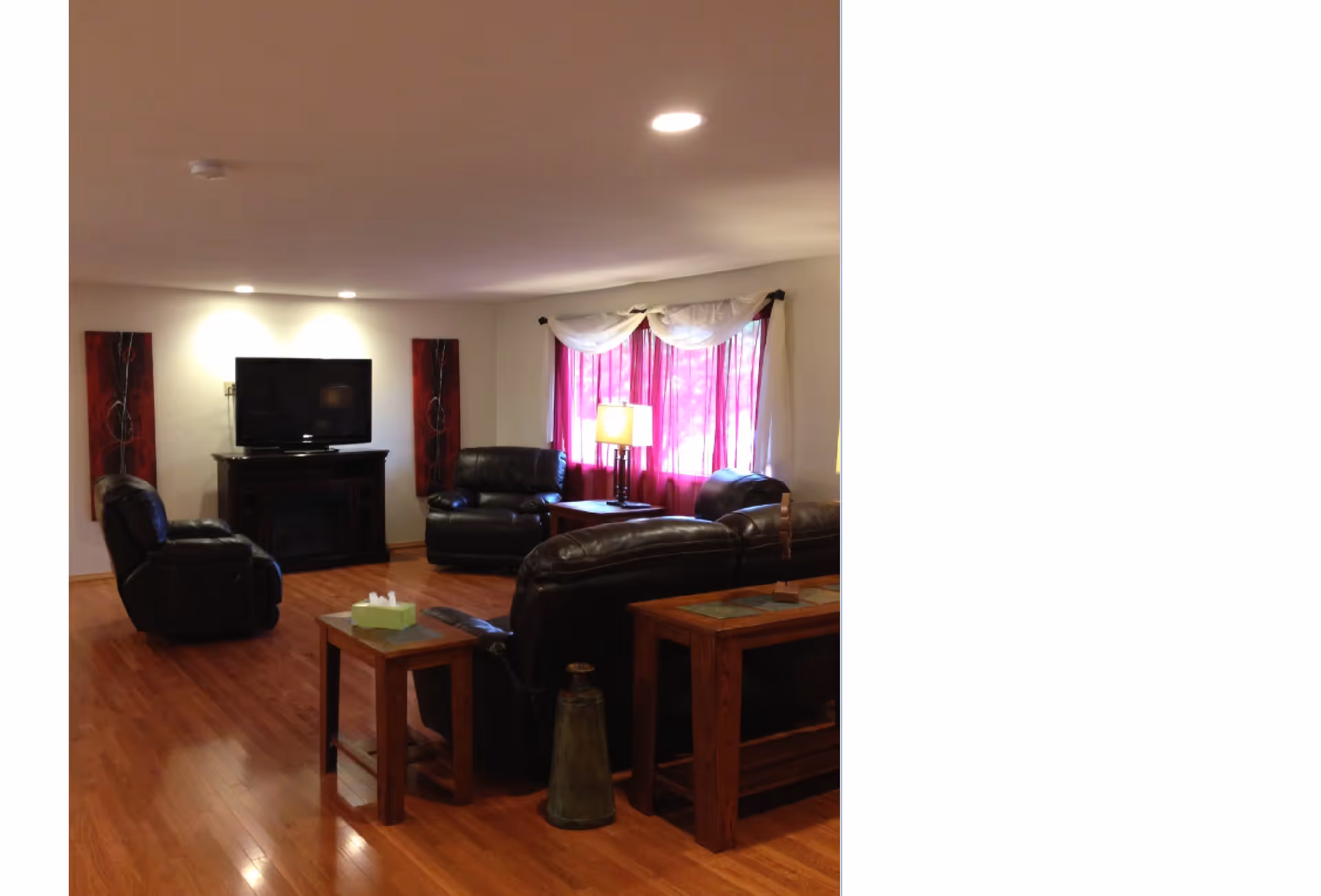 A cozy living room with wooden flooring, featuring several black leather armchairs and sofas arranged around a TV mounted on a dark wooden stand. The room is softly lit by ceiling lights and a table lamp near a window with purple curtains and white valances. There are two red decorative wall panels on either side of the TV.
