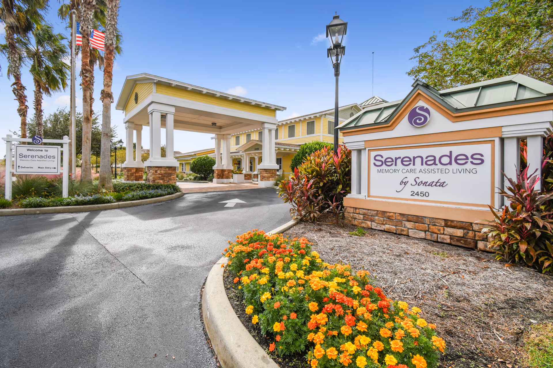 Entrance to Serenades Memory Care Assisted Living by Sonata, featuring a covered driveway, landscaped flower beds with orange and yellow flowers, palm trees, and clear blue sky.