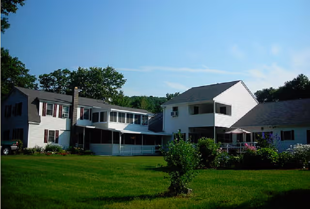 Exterior view of a large white multi-section building with dark roof shingles, surrounded by green grass and trees under a clear blue sky.