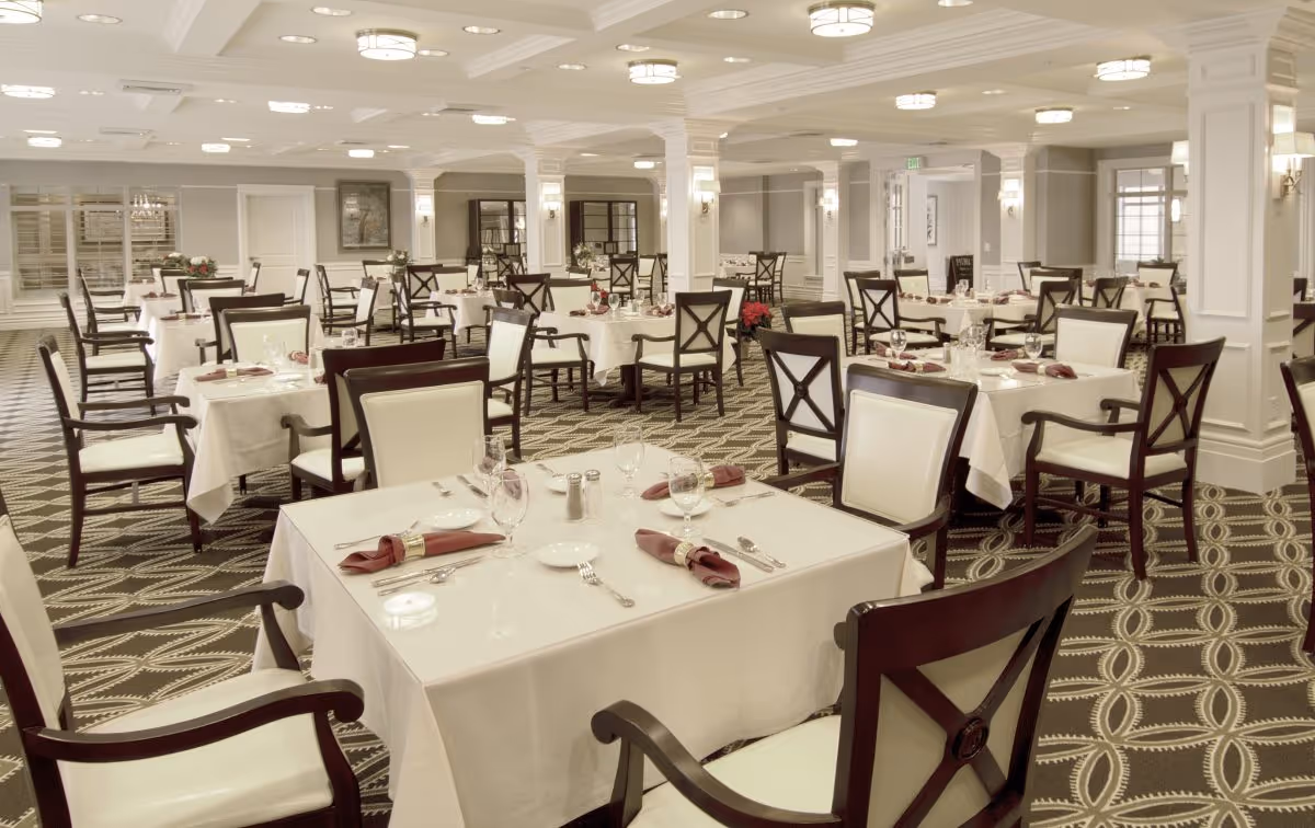 A spacious and elegant dining room with multiple tables covered in white tablecloths, each set with plates, silverware, wine glasses, and folded burgundy napkins. The room features patterned carpet, white walls with decorative columns, and soft ceiling lighting.
