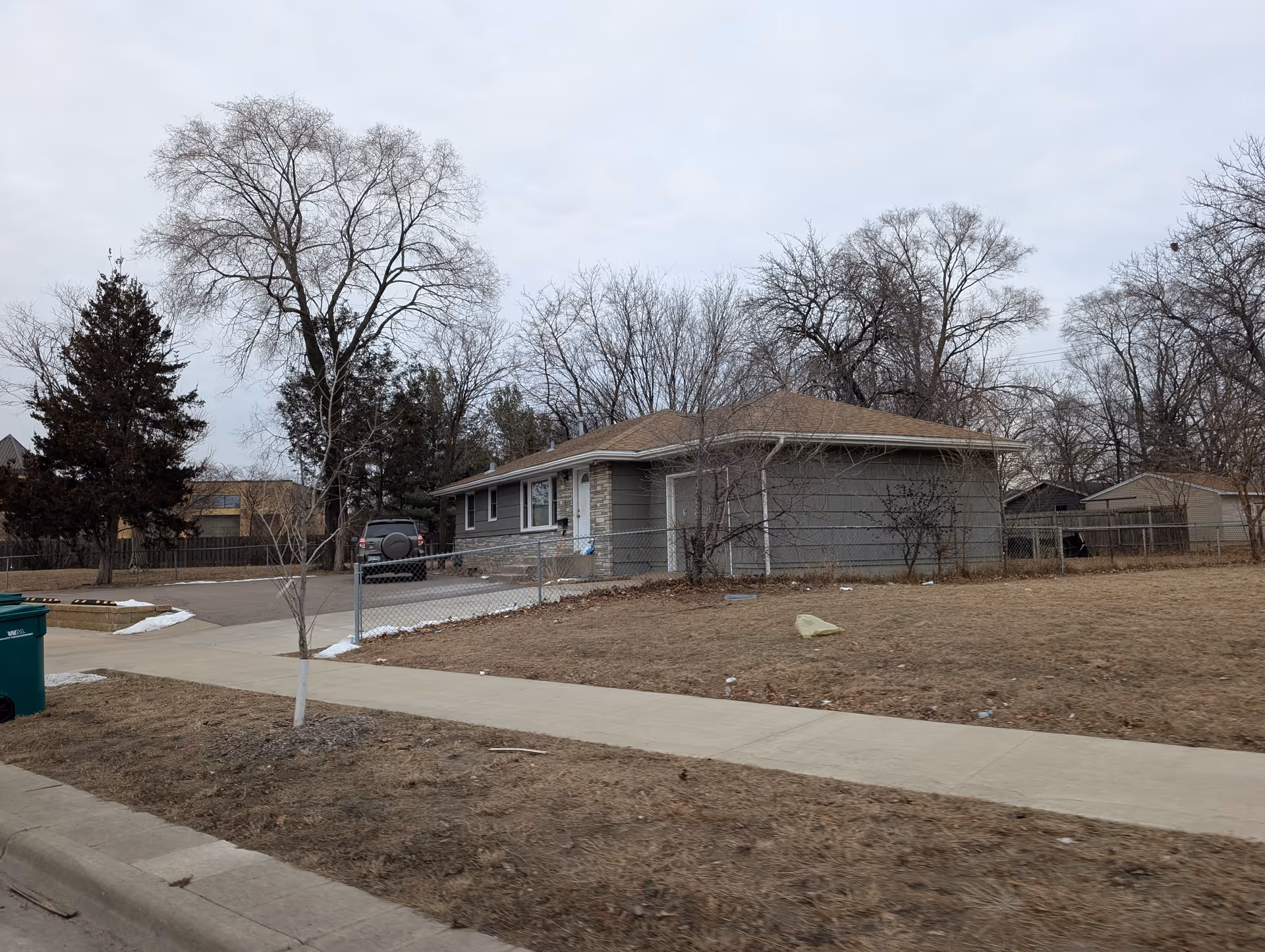 Single-story residential building with a brown roof and gray siding, surrounded by leafless trees and a chain-link fence. There is a sidewalk and a driveway with a parked vehicle. The ground is covered with dry grass and patches of snow under an overcast sky.