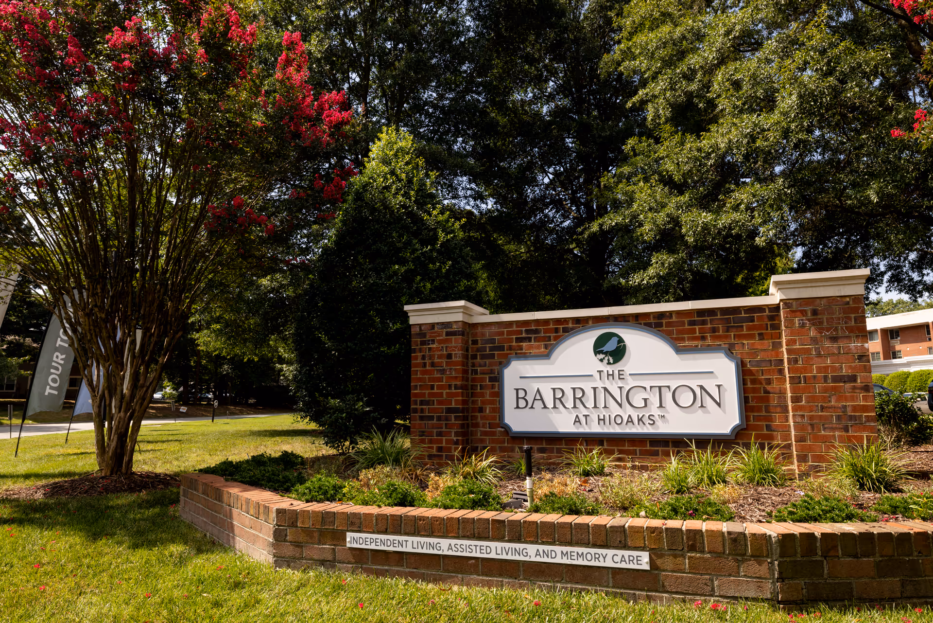 Outdoor view of a brick sign for The Barrington at Hioaks senior living facility, surrounded by green grass, trees, and flowering plants. The sign mentions independent living, assisted living, and memory care services.