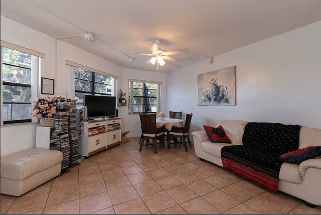 A cozy living room area with a white leather couch covered partially by a black and red blanket, a small dining table with four chairs, a TV on a stand with speakers, and a rack filled with CDs or DVDs. The room has tiled flooring, three windows with blinds, a ceiling fan with lights, and a painting of flowers on the wall.