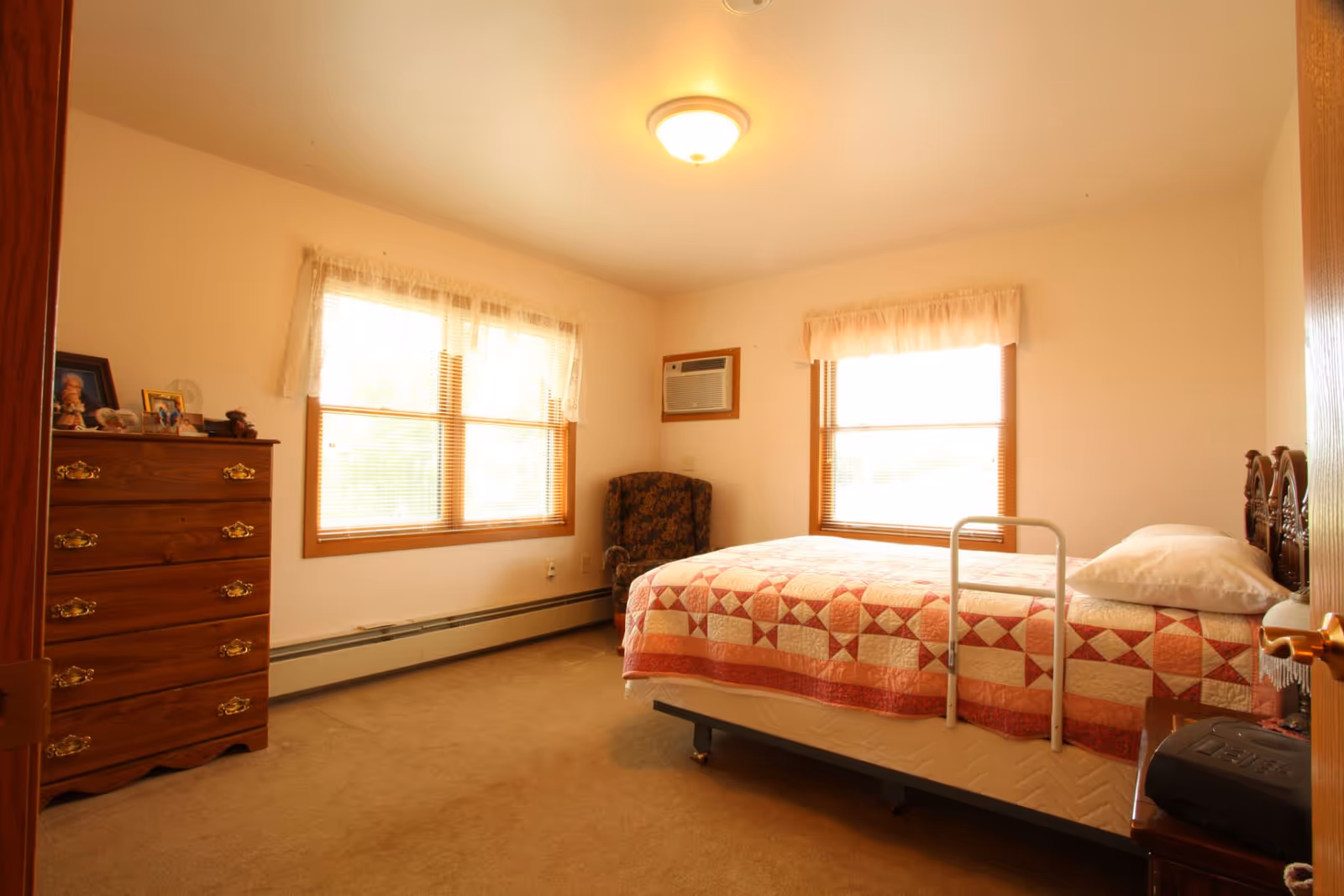 Well-lit bedroom with a single bed covered by a quilt, a wooden dresser, an armchair, and two windows.