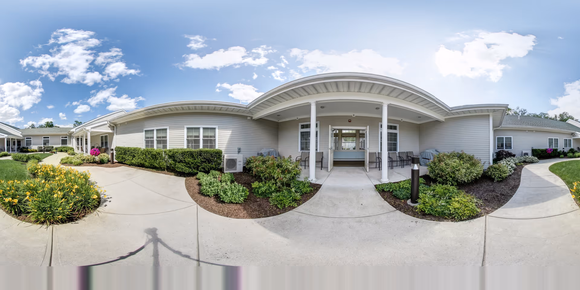 Wide panoramic view of the exterior entrance of The Lighthouse at Lincoln senior living facility, showing a single-story building with light gray siding, a covered porch supported by white columns, well-maintained landscaping with bushes and flowers, and a clear blue sky with scattered clouds.