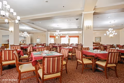 A spacious dining room with multiple wooden tables covered with red tablecloths and set with placemats and condiments. The chairs have striped upholstery in red and beige tones. The room features chandeliers hanging from the ceiling, beige walls with white trim, and large windows with curtains allowing natural light to enter.