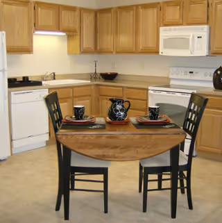 Small kitchen with oak cabinets, white appliances and a two-seat wooden dining table set with black dishware.