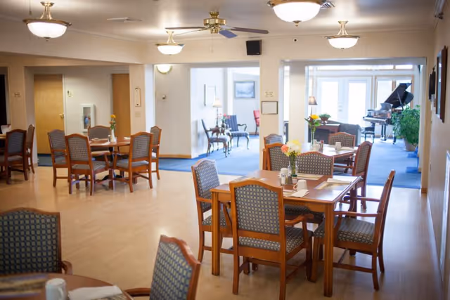 A bright and spacious dining area in an assisted living facility with several wooden tables and cushioned chairs arranged neatly. Each table has a small vase with flowers and place settings. In the background, there is a sitting area with chairs and a grand piano near large windows letting in natural light.