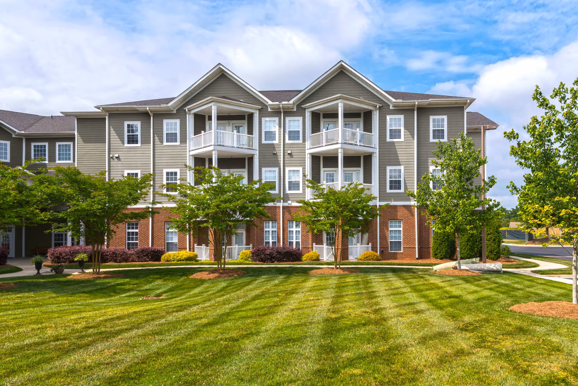 Exterior view of a three-story senior living facility building with gray siding and red brick on the lower level. The building features multiple white-trimmed windows and balconies. In front of the building is a well-maintained lawn with striped mowing patterns, several small trees, and landscaped bushes under a partly cloudy sky.