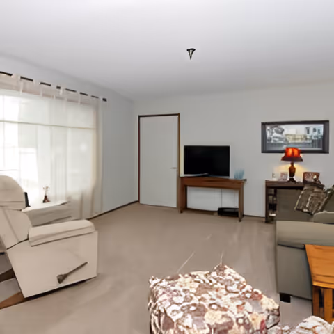 A living room with a beige recliner chair near a large window with sheer curtains, a patterned ottoman in the foreground, a wooden TV stand with a flat-screen TV, a side table with a red lamp, and a gray sofa with cushions.