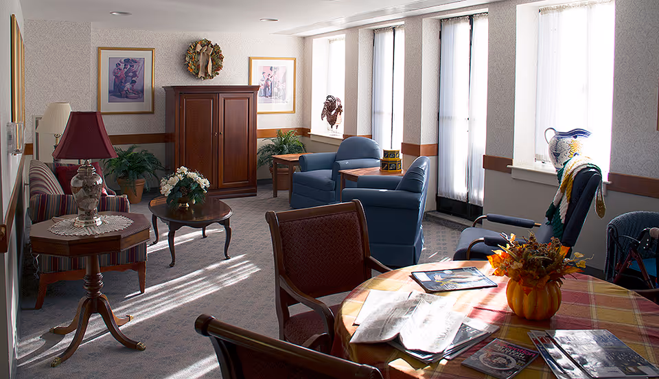 A cozy living room area with several chairs and tables. There is a striped sofa with a lamp on a side table, two blue armchairs with a small table between them, and a round table covered with a plaid tablecloth holding magazines and a decorative vase with flowers. The room has large windows with sheer curtains letting in natural light, framed artwork on the walls, and a wooden cabinet against the far wall. The carpet is light-colored and the overall atmosphere is warm and inviting.